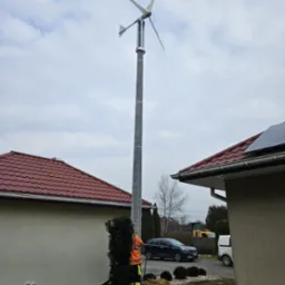 Montaż turbiny wiatrowej przy domu jednorodzinnym. Widoczny wysoki maszt z turbiną, pracownik w odblaskowej kamizelce oraz fragment dachu z panelem słonecznym.