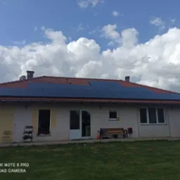 Dom jednorodzinny z panelami słonecznymi na czerwonym dachu, widok z ogrodu w słoneczny dzień z chmurami. Część elewacji w trakcie malowania.