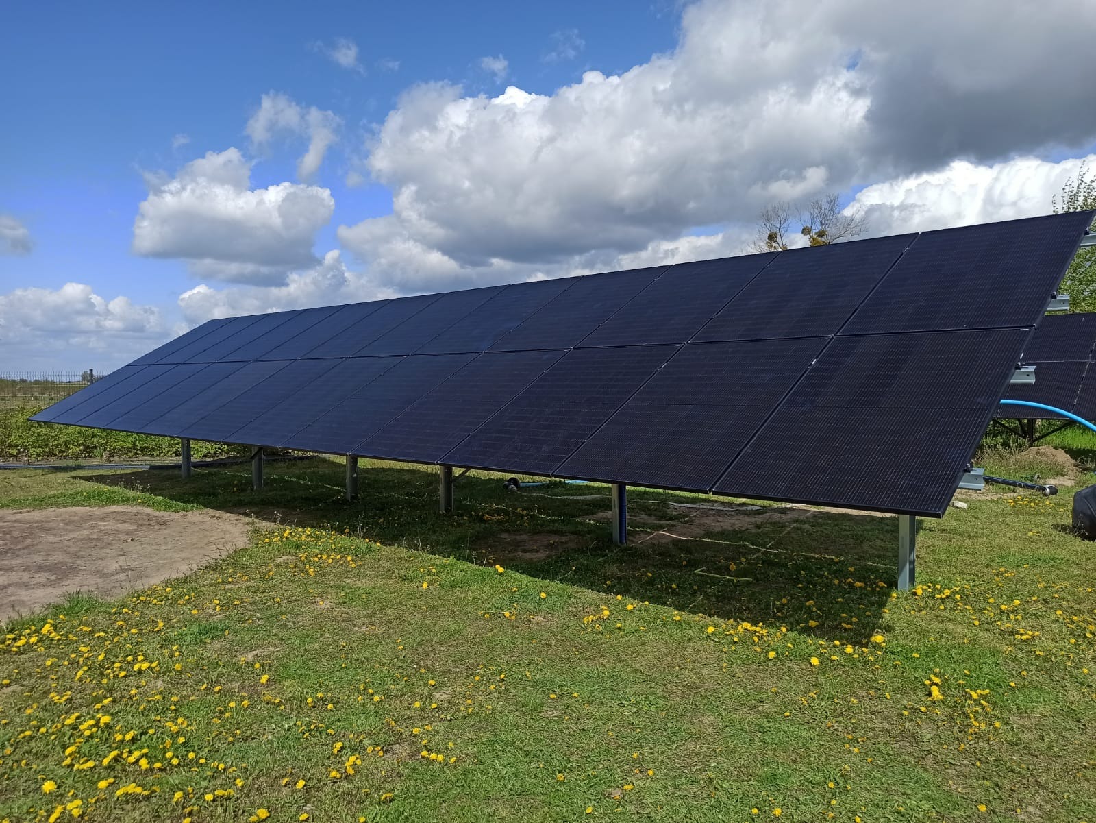 Rząd paneli słonecznych na łące z mleczami pod błękitnym niebem. Widoczne metalowe podpory i fragment instalacji. Kąt ujęcia z dołu, podkreślający rozmiar instalacji.