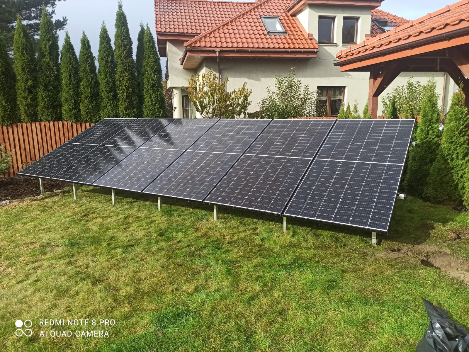 Panele słoneczne na trawniku przed domem jednorodzinnym. Moduły ustawione na metalowych wspornikach, widoczne drzewa iglaste i część elewacji budynku.