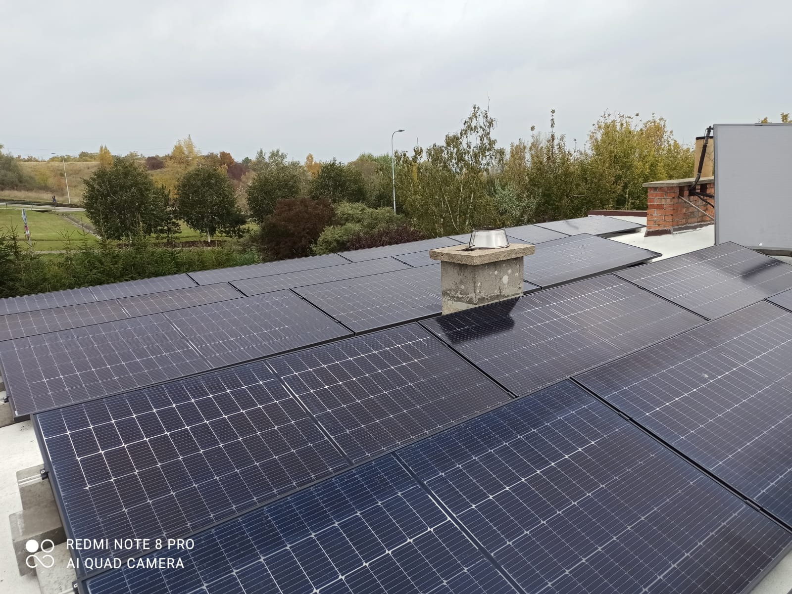 Panele słoneczne na dachu budynku w Ornecie, widok z góry. Obok komin z metalowym daszkiem. W tle drzewa i pochmurne niebo.