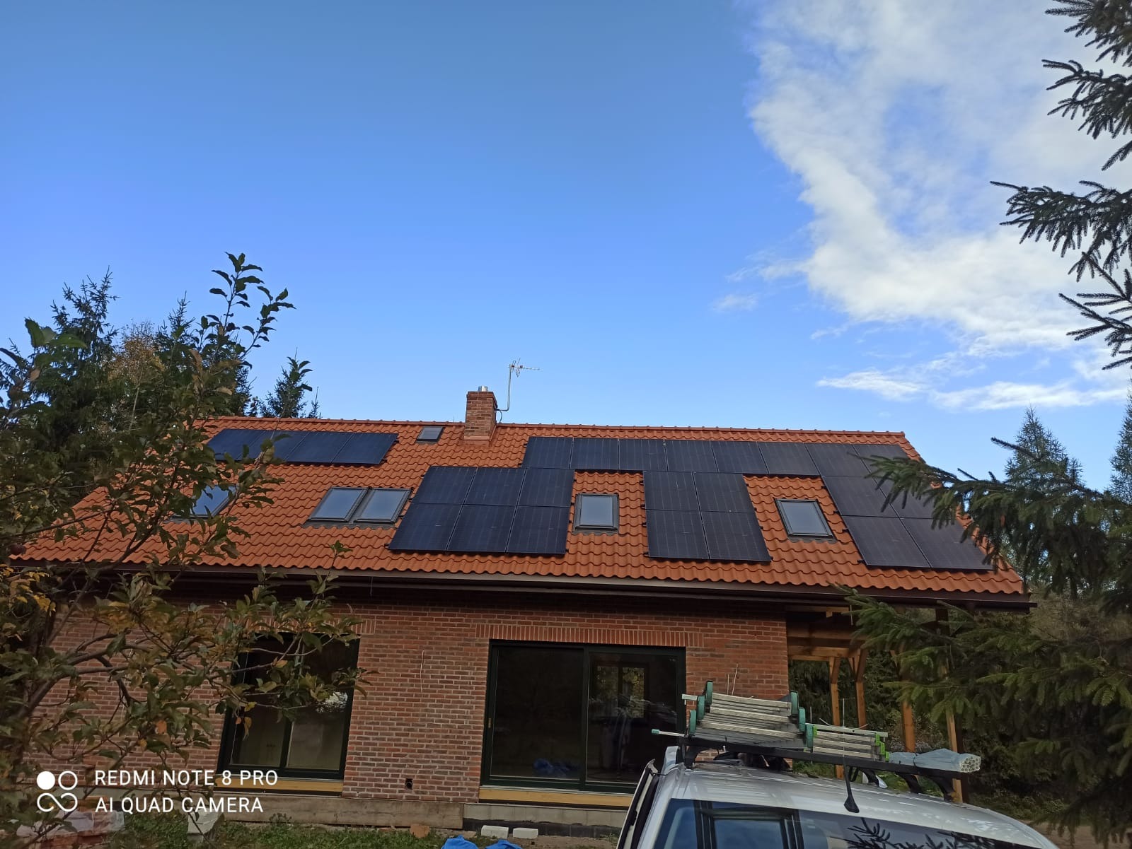 Czerwony dach z panelami słonecznymi i oknami dachowymi na ceglanym domu jednorodzinnym w słoneczny dzień. Na dachu samochód z drabiną.