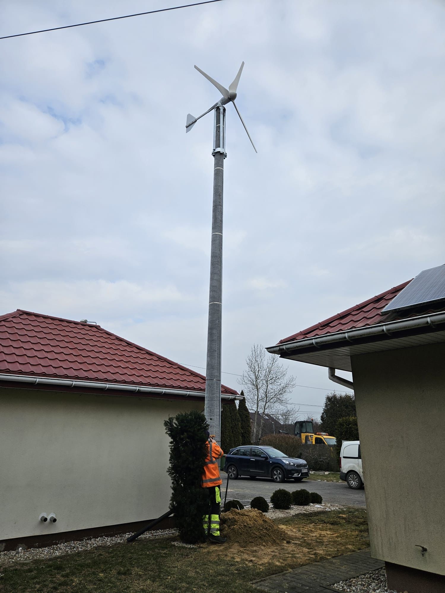 Montaż turbiny wiatrowej przy domu jednorodzinnym. Widoczny wysoki maszt z turbiną, pracownik w odblaskowej kamizelce oraz fragment dachu z panelem słonecznym.