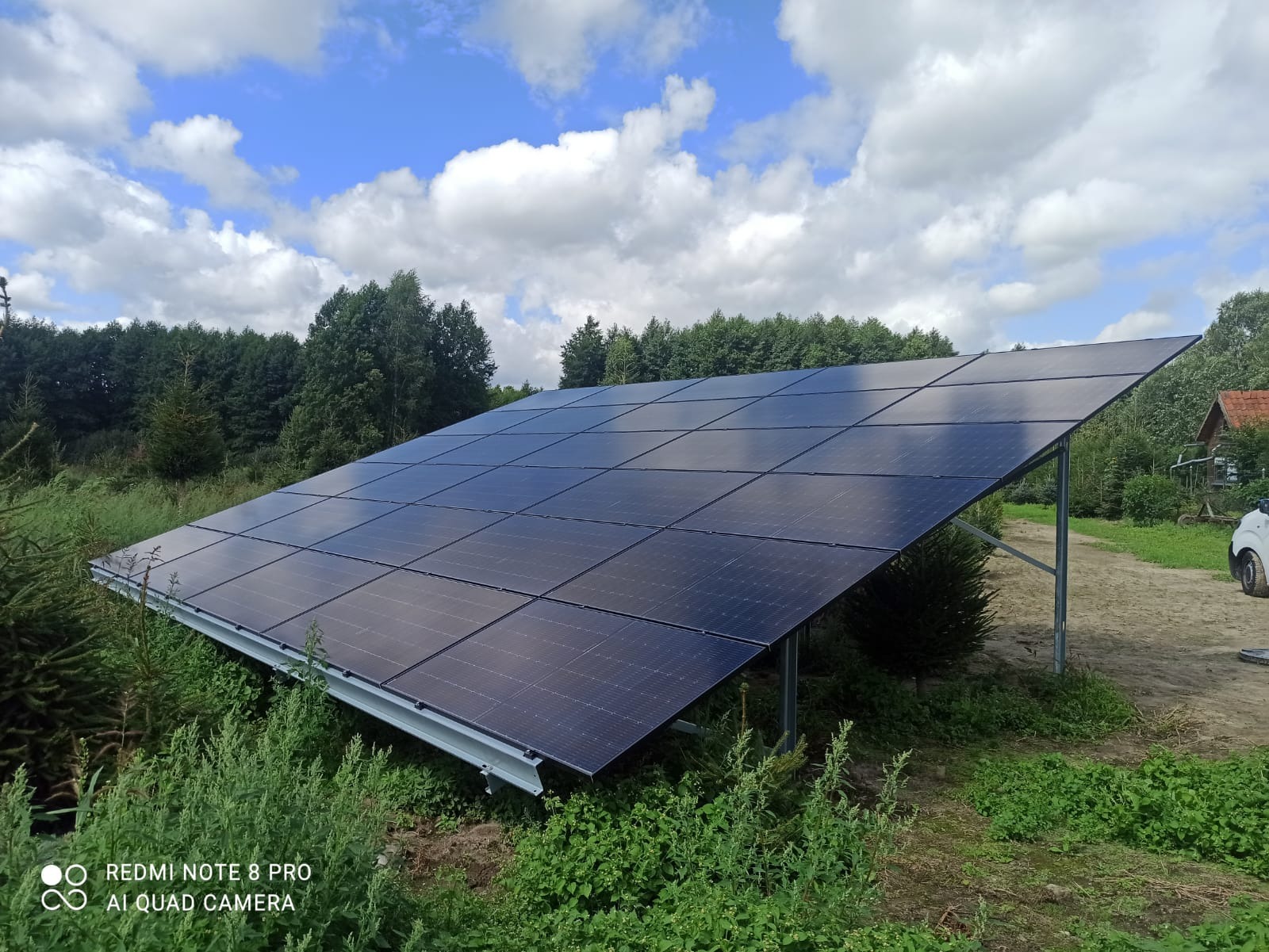 Panel słoneczny zamontowany na konstrukcji w terenie zielonym, z widocznym niebem i chmurami. Ujęcie z dołu, podkreślające wielkość instalacji.