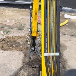 Grzegorz Olender - Żółta minikoparka Interbauer w trakcie pracy, widoczne przewody hydrauliczne zabezpieczone metalowymi obejmami. Ślady wykopu w ziemi, fragment budynku w tle.