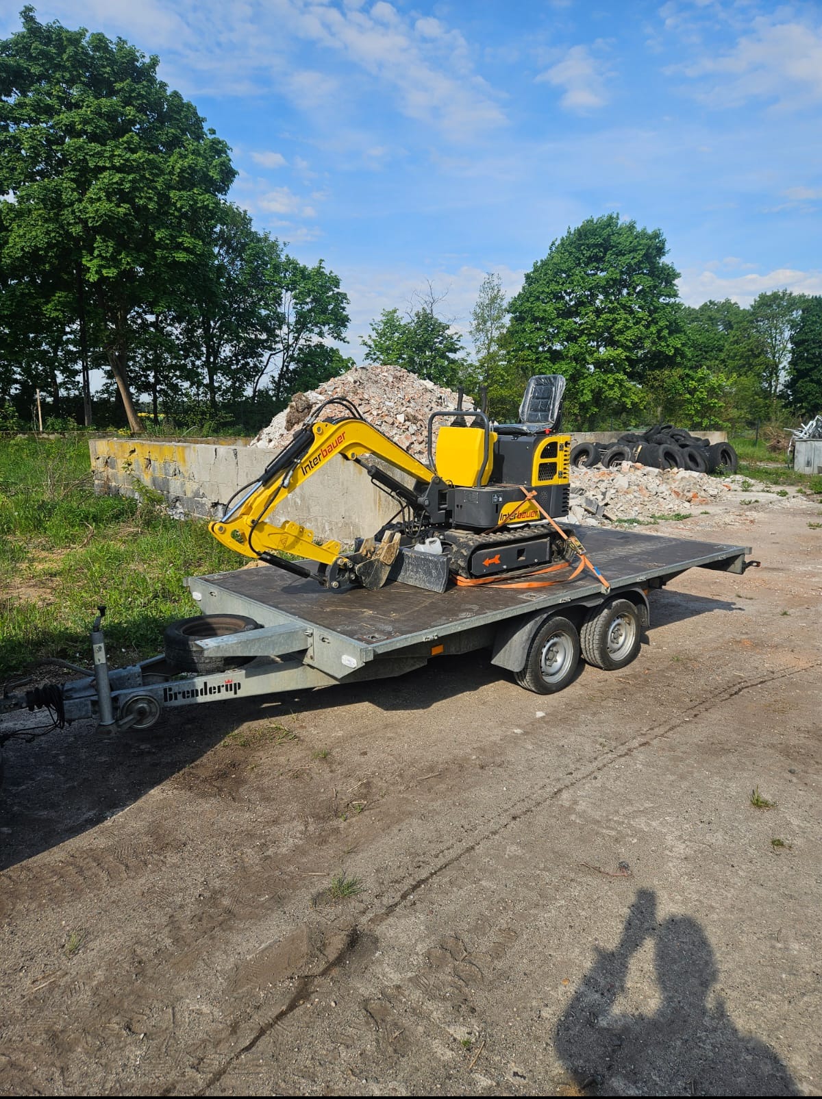 Żółta mini koparka Interbauer na przyczepie, zabezpieczona pasami. W tle gruz i opony, częściowo zburzona konstrukcja. Słoneczny dzień.