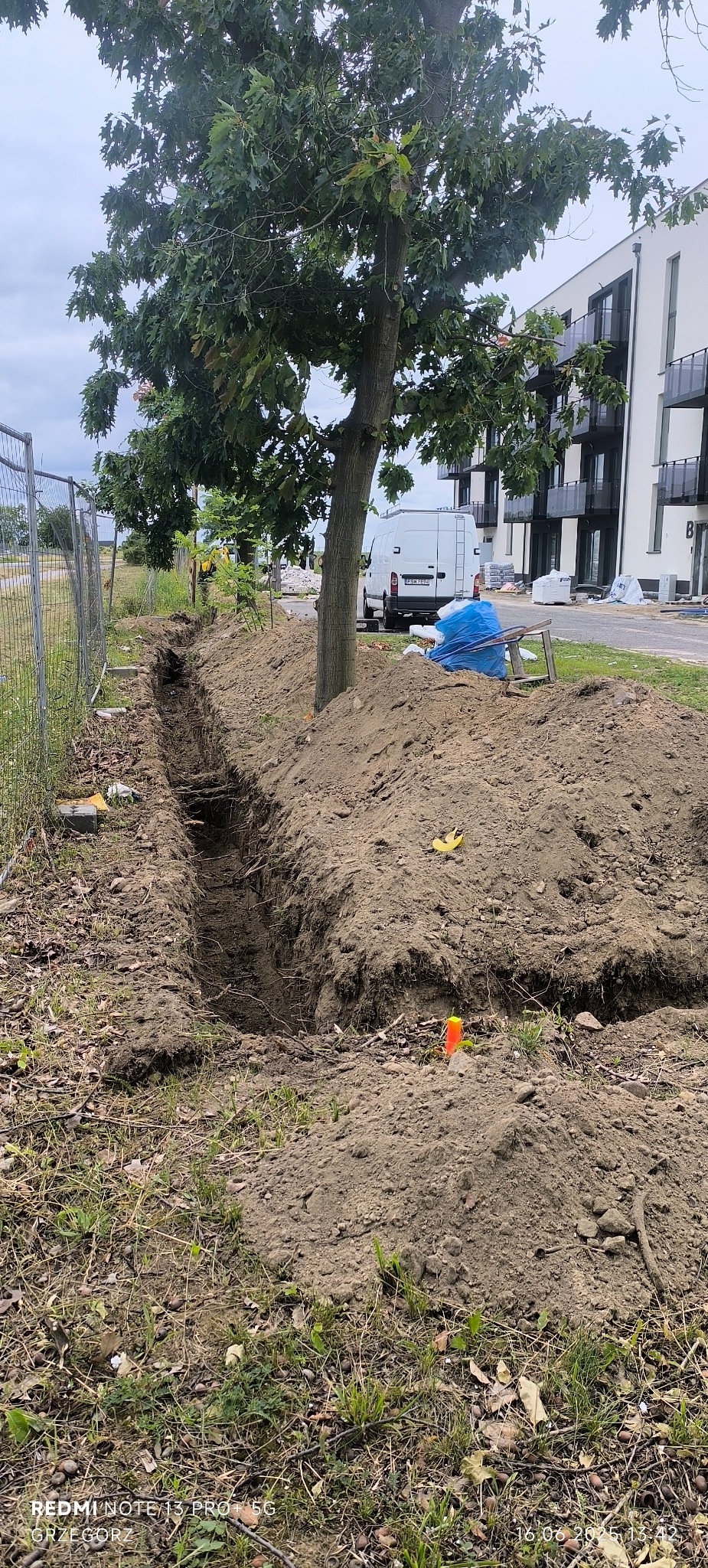 Wykopany wąski rów w ziemi, obok drzewo i pryzma ziemi. W tle budynek mieszkalny i zaparkowany biały bus. Pomarańczowy słupek w ziemi.