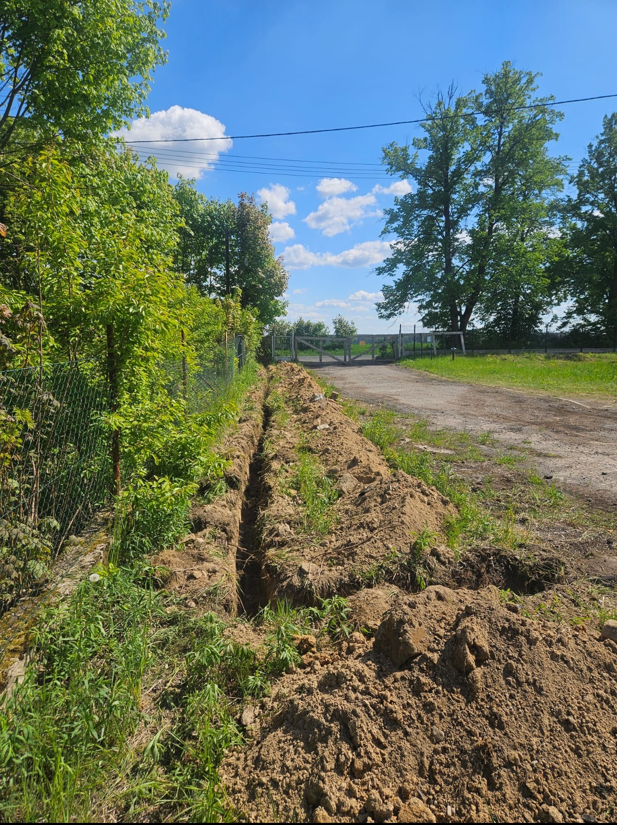 Wykopany wąski rów w ziemi, prawdopodobnie pod instalacje, biegnący wzdłuż ogrodzenia z siatki, z widokiem na polną drogę i zieleń w tle. Słoneczny dzień.