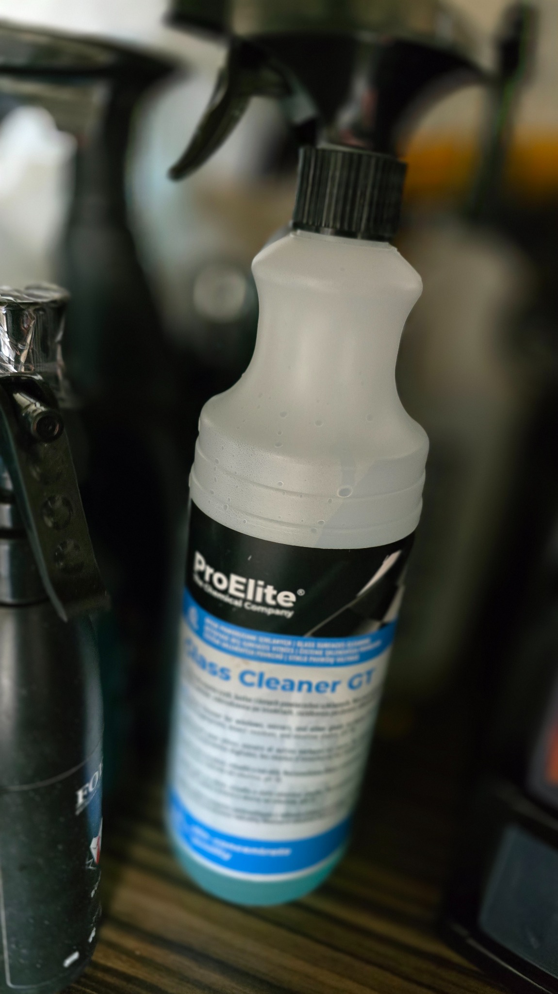 Butelka środka czyszczącego ProElite Glass Cleaner GT z rozpylaczem, stojąca na drewnianej powierzchni, obok innych butelek z chemią. Widoczne krople na butelce.