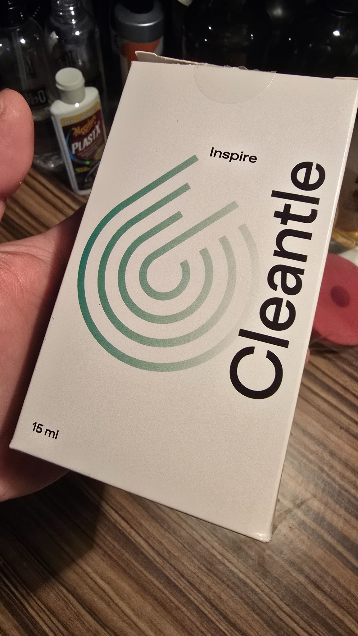 Białe pudełko produktu Cleantle Inspire 15 ml, trzymane w dłoni, z zielonym logo i czarnym napisem na tle drewnianego blatu i innych butelek.