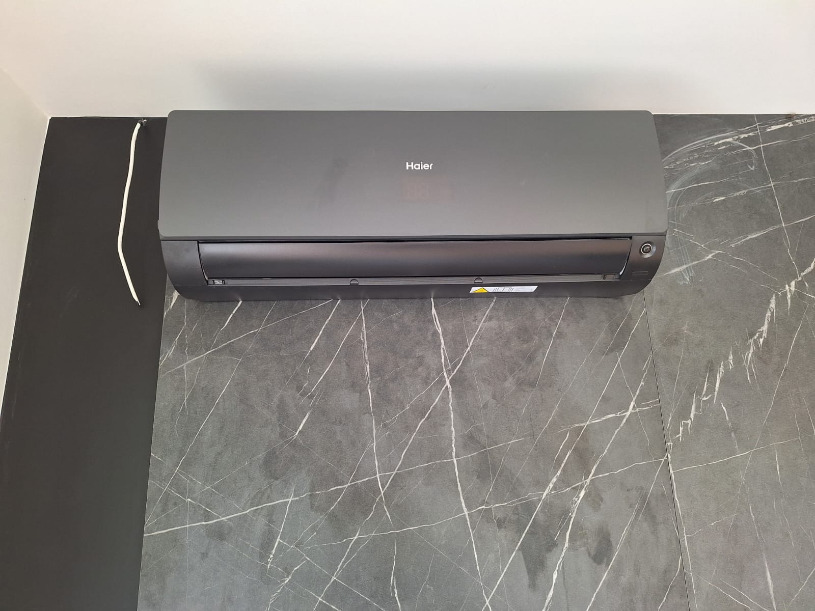 Szary klimatyzator Haier na ścianie z szarego marmuru z białymi żyłami. Widoczny biały kabel. Nowoczesne wnętrze.