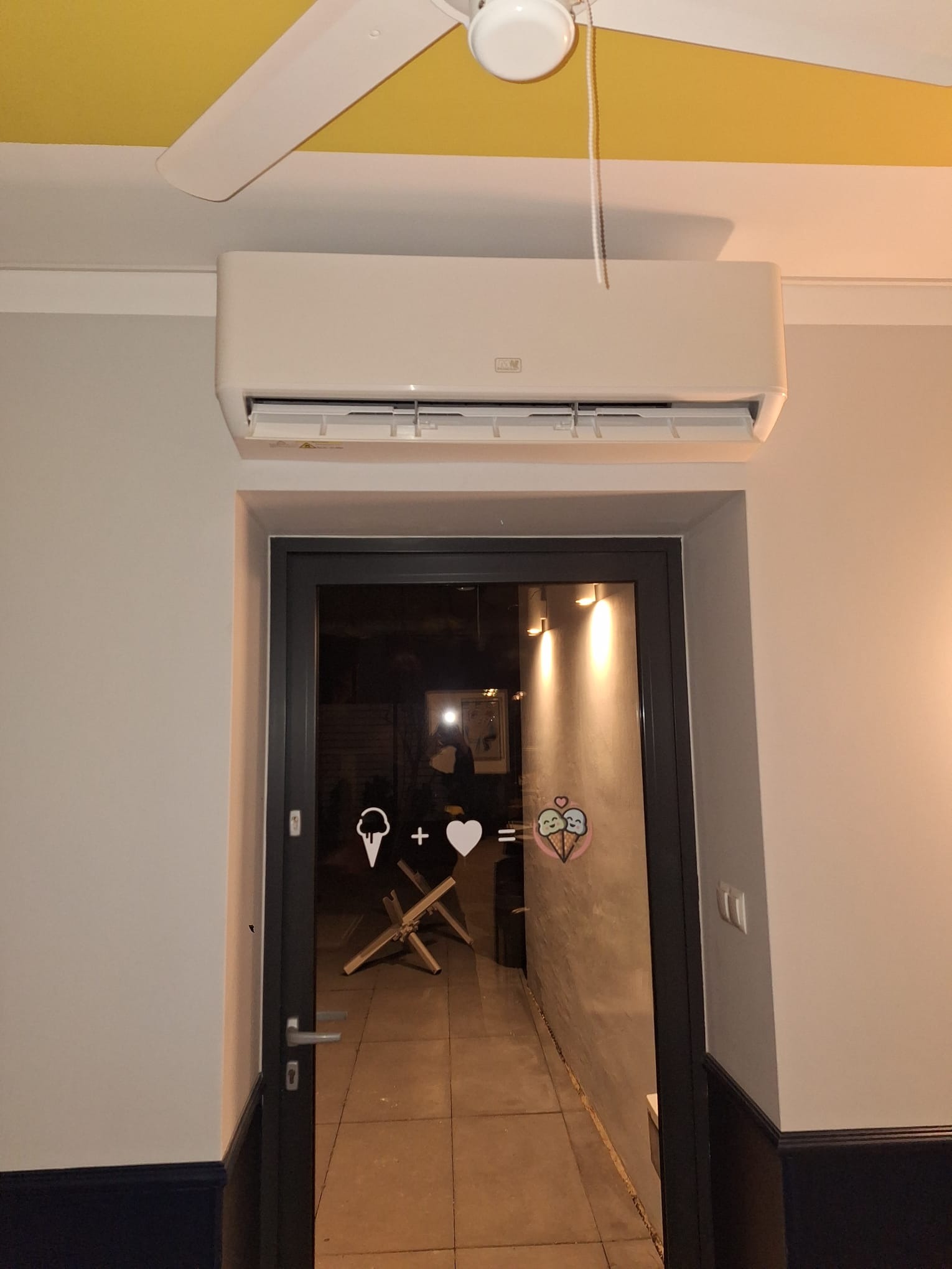 Biała klimatyzacja zamontowana nad wejściem do lokalu z naklejką lodów na szklanych drzwiach. Widoczny fragment sufitu i wentylator.