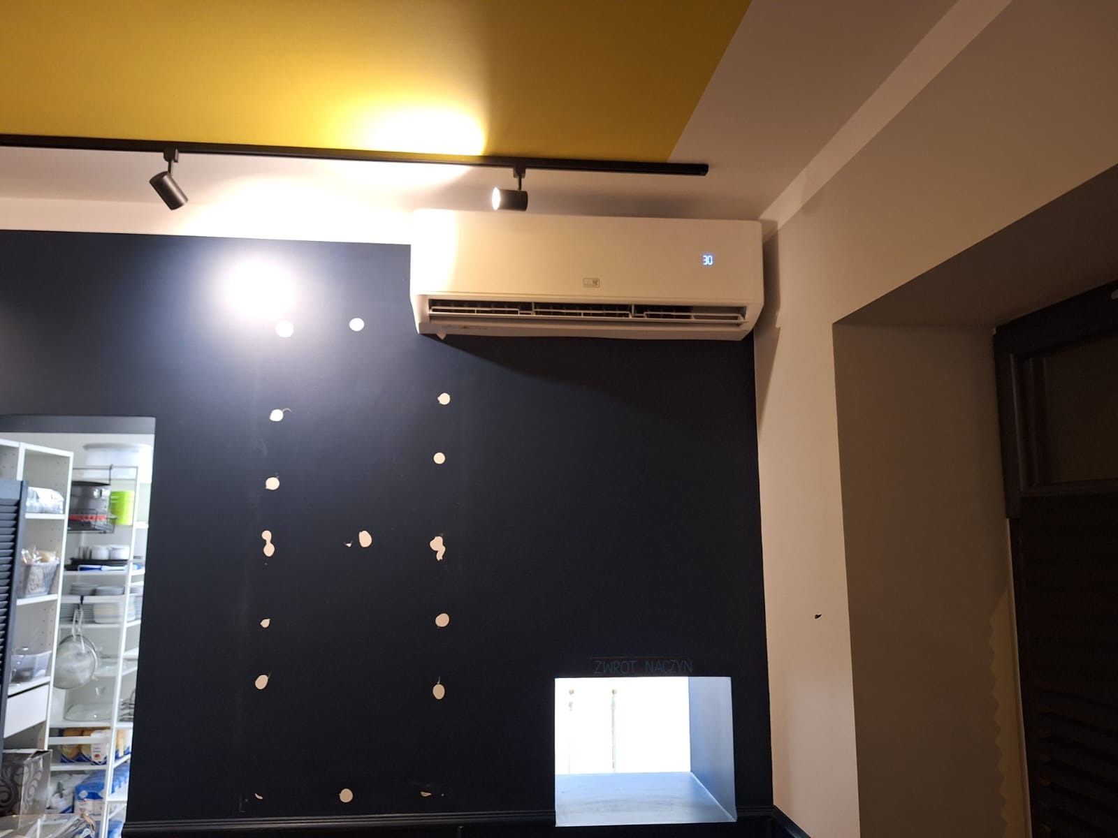 Biały klimatyzator ścienny zamontowany w pomieszczeniu z ciemną ścianą i żółtym sufitem, widoczne ślady po montażu na ścianie.