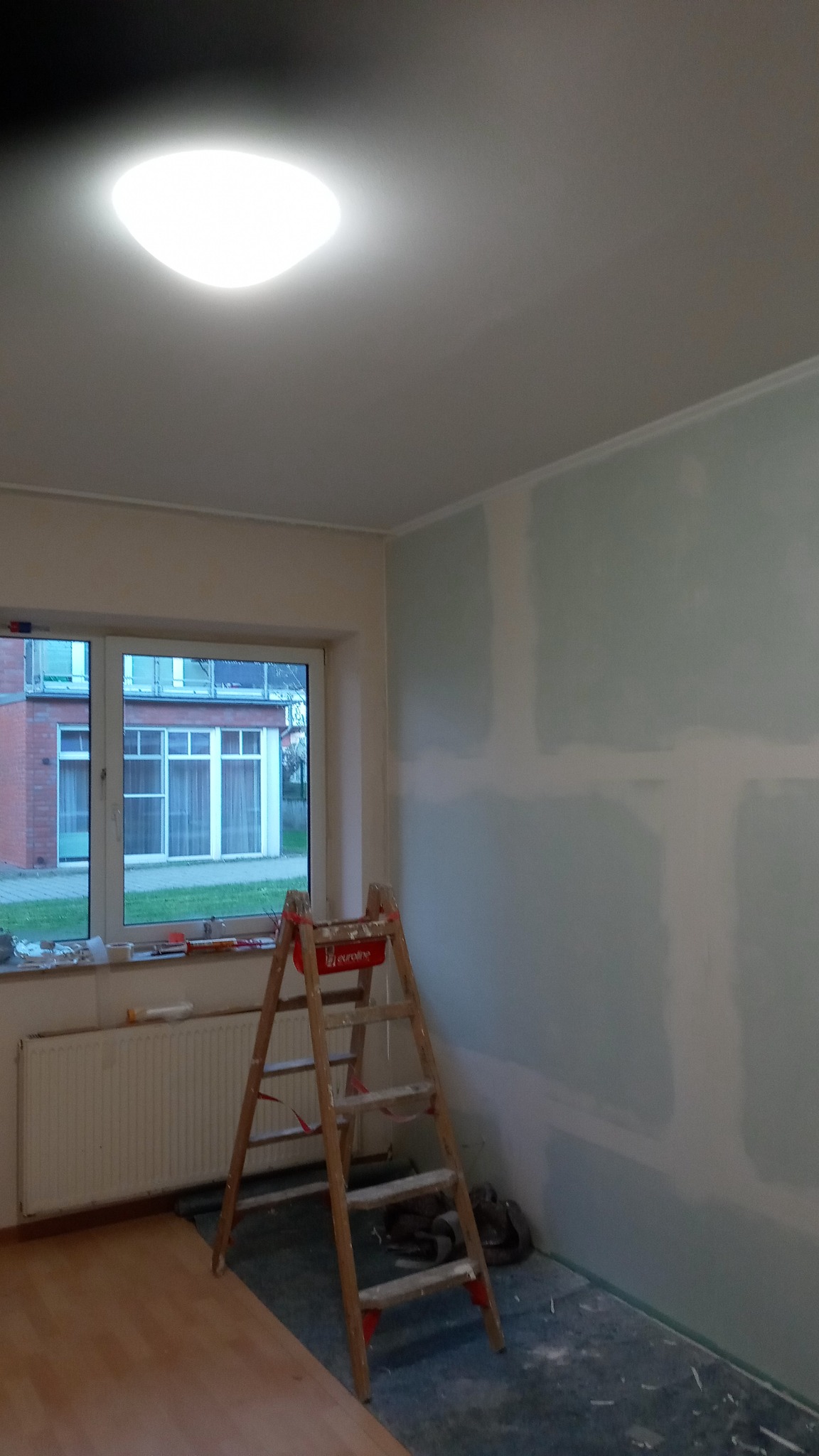 Ściana po szpachlowaniu w Cekcyn, widoczna drabina i okno. Przygotowanie do malowania. Ściana w jasnym odcieniu z widocznymi łączeniami płyt gipsowo-kartonowych.