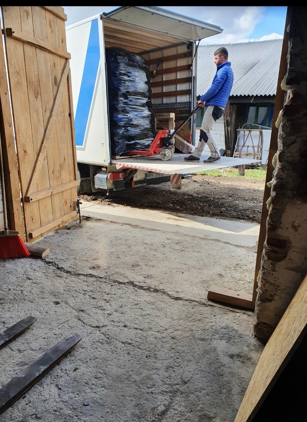 Pracownik z wózkiem paletowym wyładowuje zapakowane towary z ciężarówki. Widoczne drewniane drzwi i fragment podwórza.