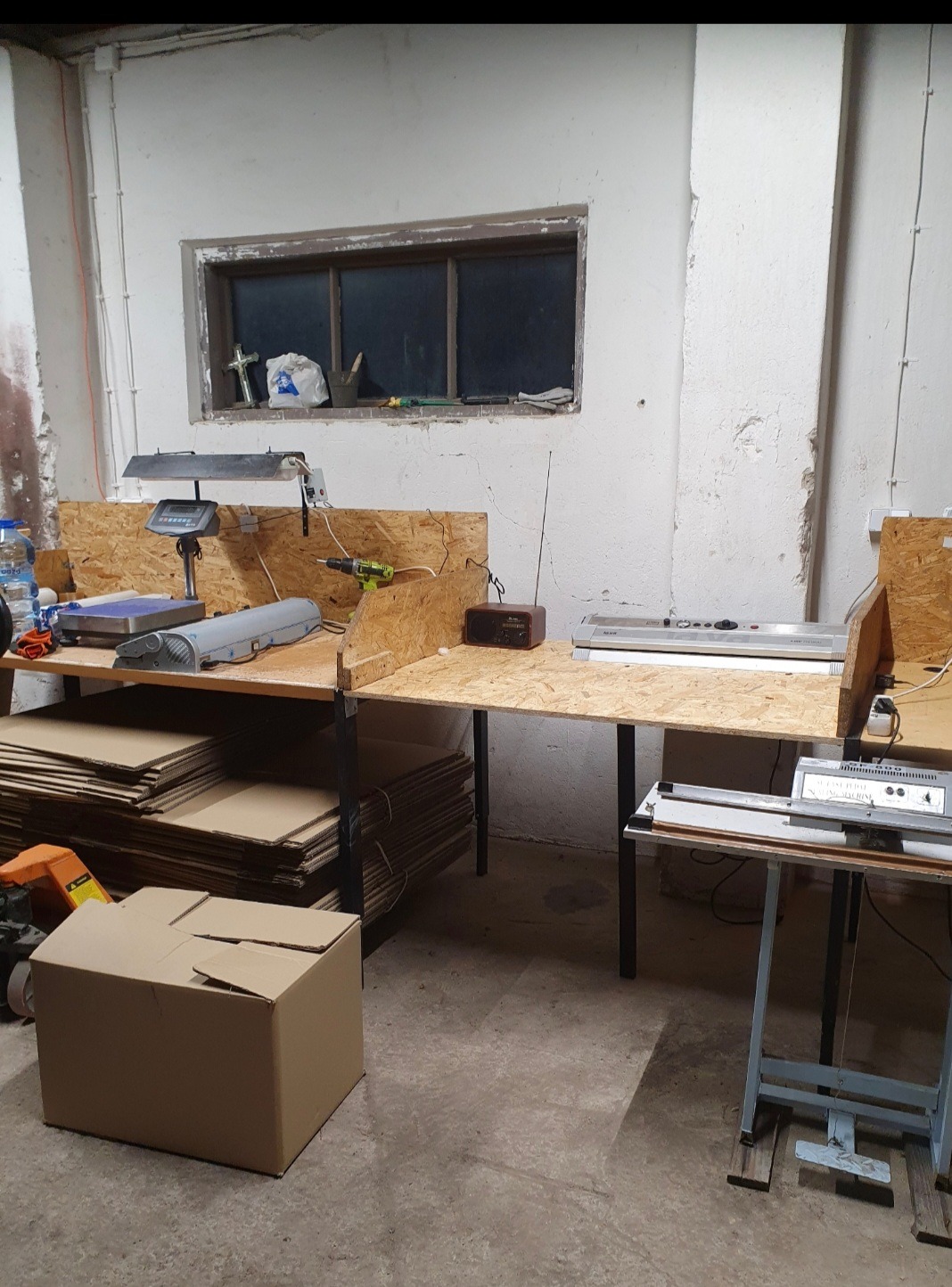 Stanowisko do pakowania: stoły z płyt OSB, waga, maszyna do pakowania, kartony. Widoczne narzędzia i materiały eksploatacyjne. Surowe wnętrze magazynu.