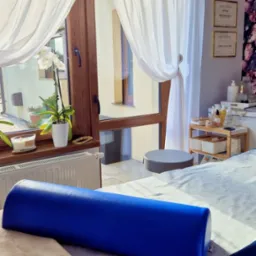 Jasne wnętrze gabinetu z łóżkiem do zabiegów, niebieskim wałkiem i oknem z białymi firanami. Na ścianie wiszą certyfikaty.