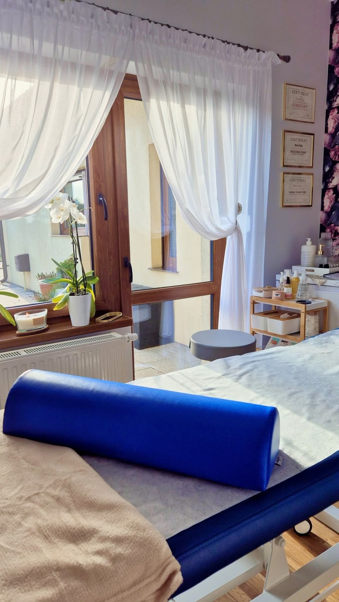 Jasne wnętrze gabinetu z łóżkiem do zabiegów, niebieskim wałkiem i oknem z białymi firanami. Na ścianie wiszą certyfikaty.