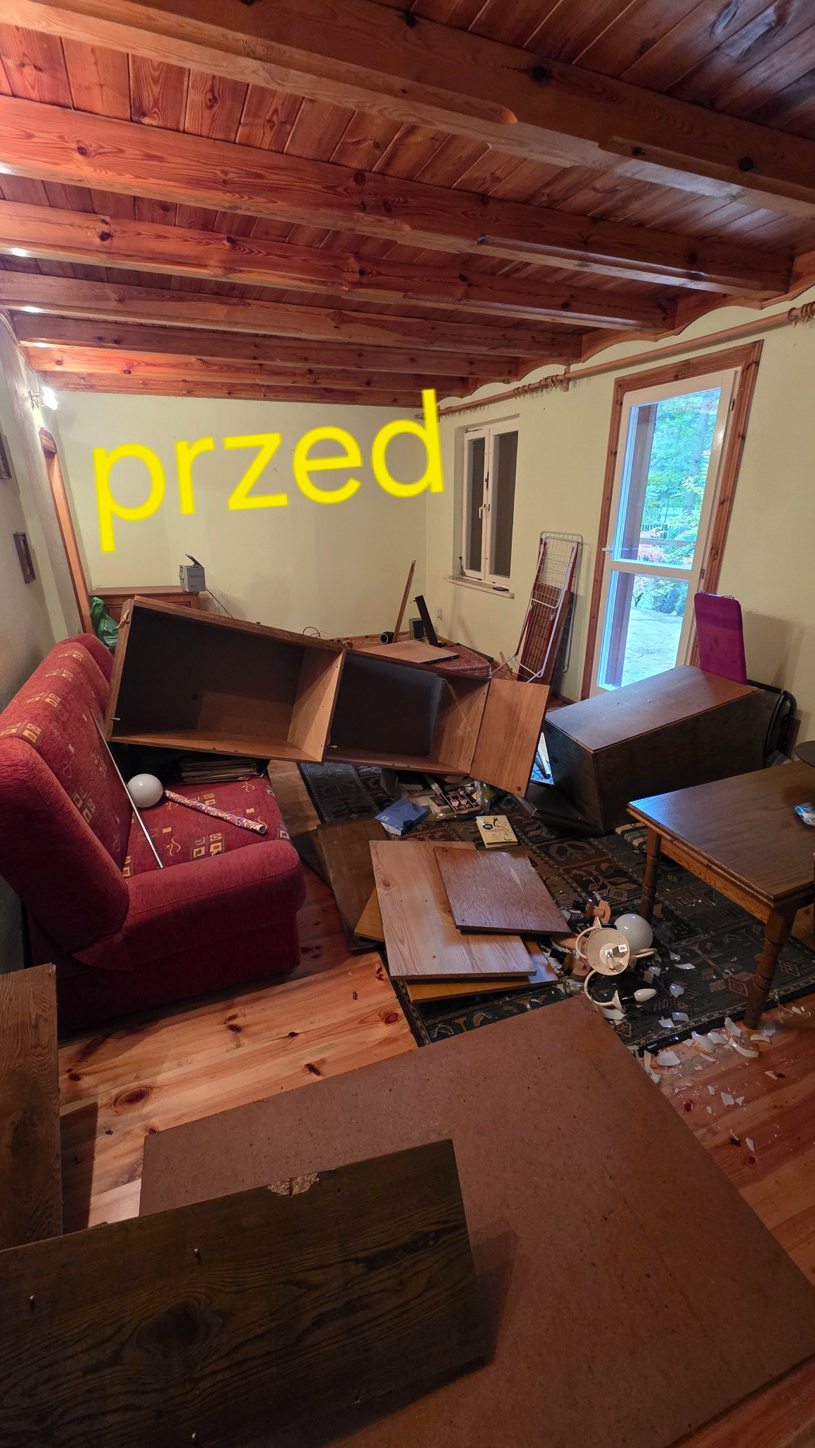 Pokój przed opróżnianiem: przewrócone meble, porozrzucane przedmioty, widoczny bałagan na podłodze i dywanie. Drewniany sufit z belkami. Ujęcie z perspektywy.