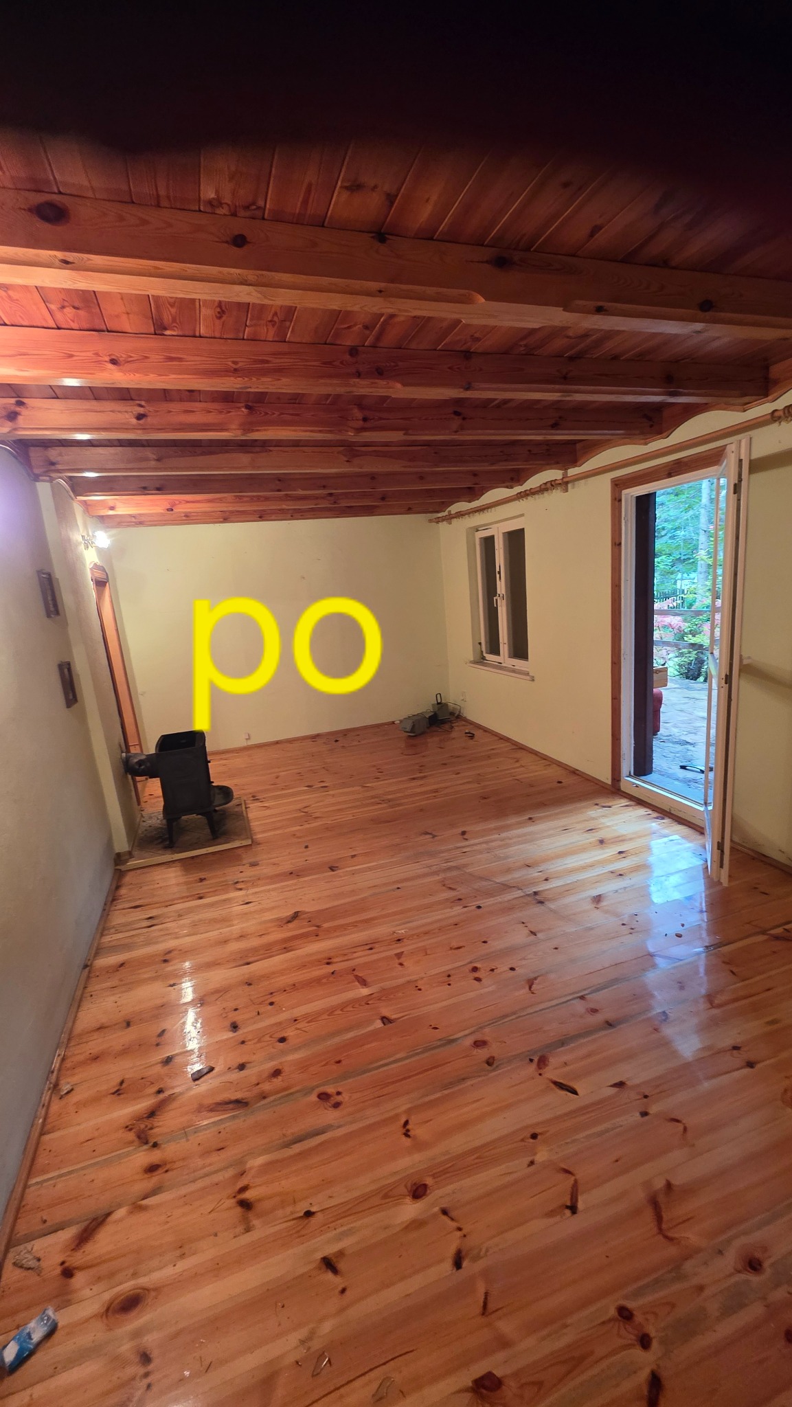 Jasne, odnowione drewniane podłogi i sufit w pustym pokoju z piecem i otwartymi drzwiami. Widoczny napis 'po'.