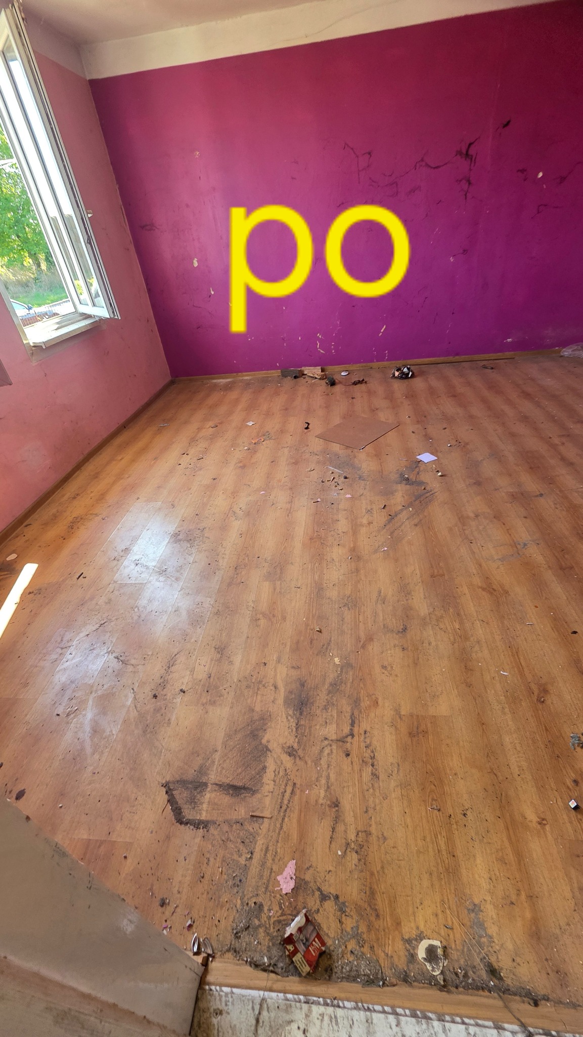 Pokój po remoncie z fioletową ścianą i drewnianą podłogą, na której widoczne są resztki gruzu i śmieci. Na ścianie napis 'po'.