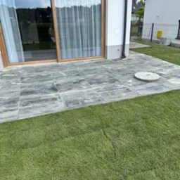 Nowoczesny taras z kostki brukowej o szarym odcieniu, przylegający do budynku z dużym oknem przesuwnym i świeżo założonym trawnikiem. Widoczny właz kanalizacyjny.