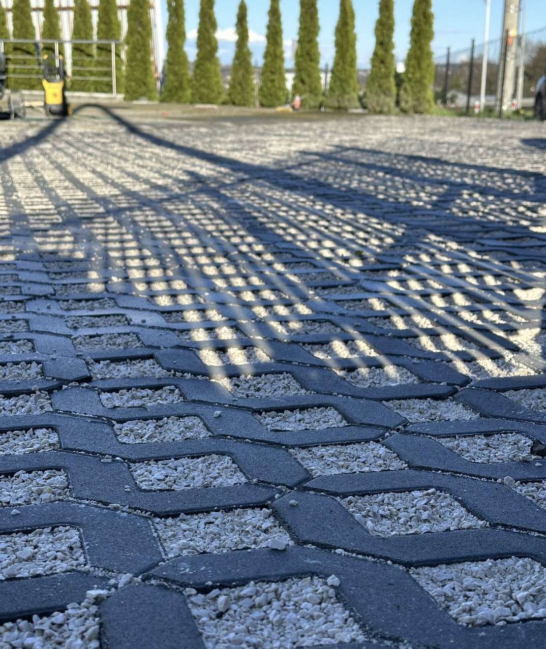 Szara ażurowa kostka wypełniona białym grysem na parkingu. W tle rząd tuj i błękitne niebo. Widoczne cienie drzew. Bliski plan, ostre światło.