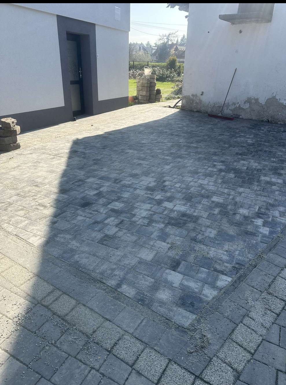 Szara kostka brukowa ułożona na posesji między budynkami, widoczne detale łączeń i faktury kostki, w tle elementy zieleni i fragment budynku.