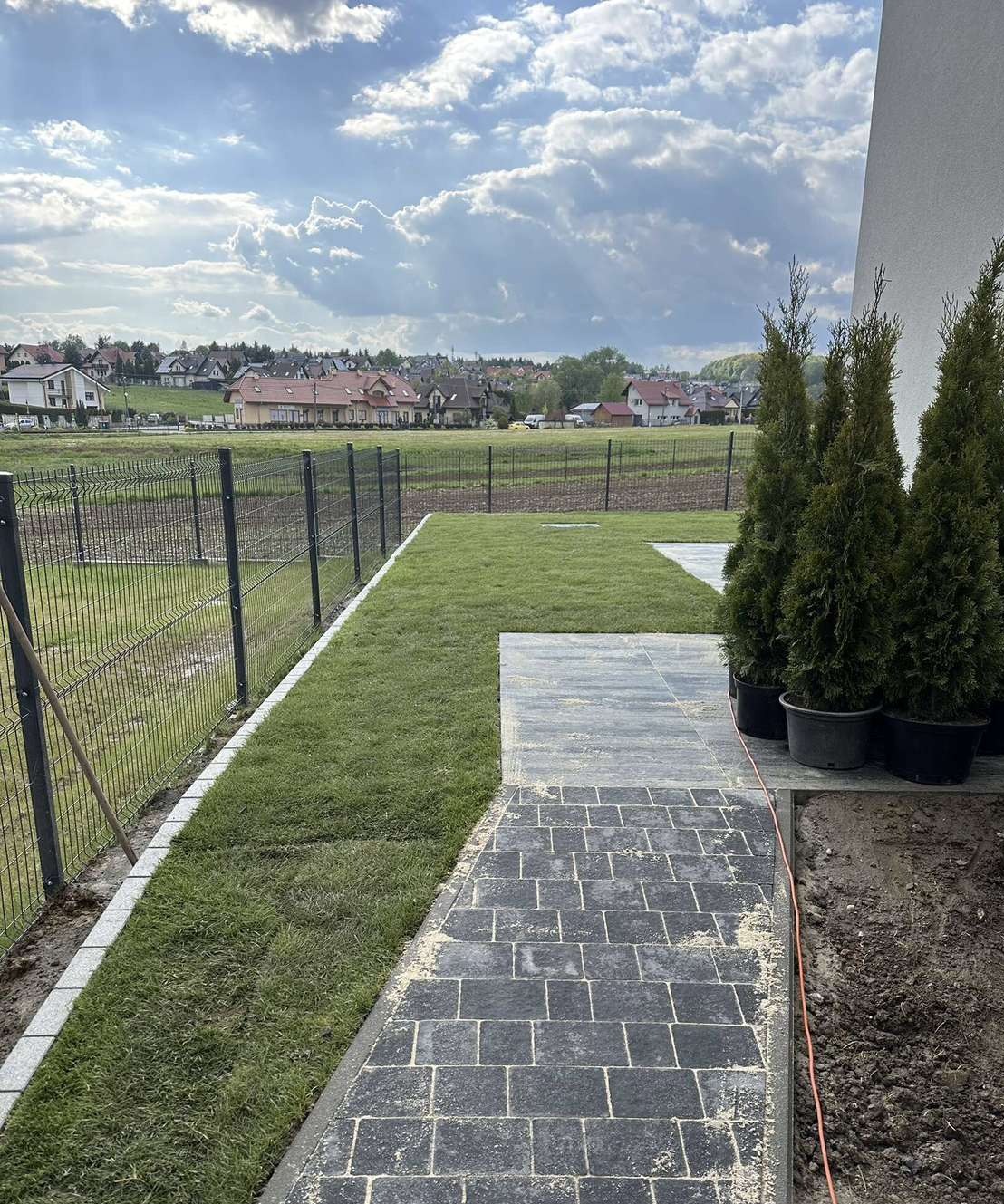 Ścieżka z grafitowej kostki brukowej, obrzeża trawnika z jasnych płyt, metalowe ogrodzenie panelowe na tle zabudowy jednorodzinnej i pochmurnego nieba.
