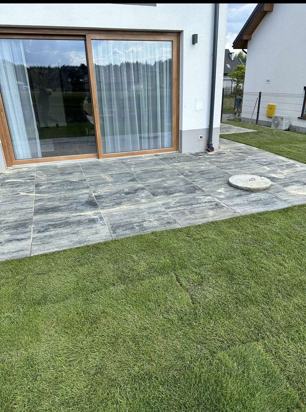 Nowoczesny taras z kostki brukowej o szarym odcieniu, przylegający do budynku z dużym oknem przesuwnym i świeżo założonym trawnikiem. Widoczny właz kanalizacyjny.