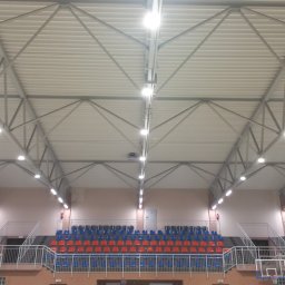 LEDINSIDE INVEST SPÓŁKA Z OGRANICZONĄ ODPOWIEDZIALNOŚCIĄ - Hala sportowa z widocznymi rzędami krzeseł i oświetleniem sufitowym. Metalowa konstrukcja dachu z równomiernie rozmieszczonymi lampami. Widok z dołu na sufit.