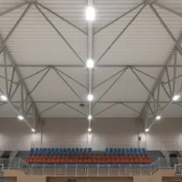 Hala sportowa z widoczną konstrukcją dachu i rzędami krzeseł. Zamontowane, równomiernie rozmieszczone lampy sufitowe oświetlają przestrzeń.
