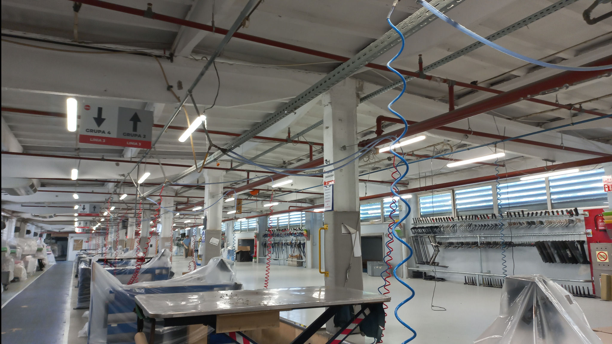 Fabryczna hala z rzędami stanowisk pracy pod rzędem lamp i instalacji podwieszonych do sufitu. Widoczne przewody, rury i oświetlenie.