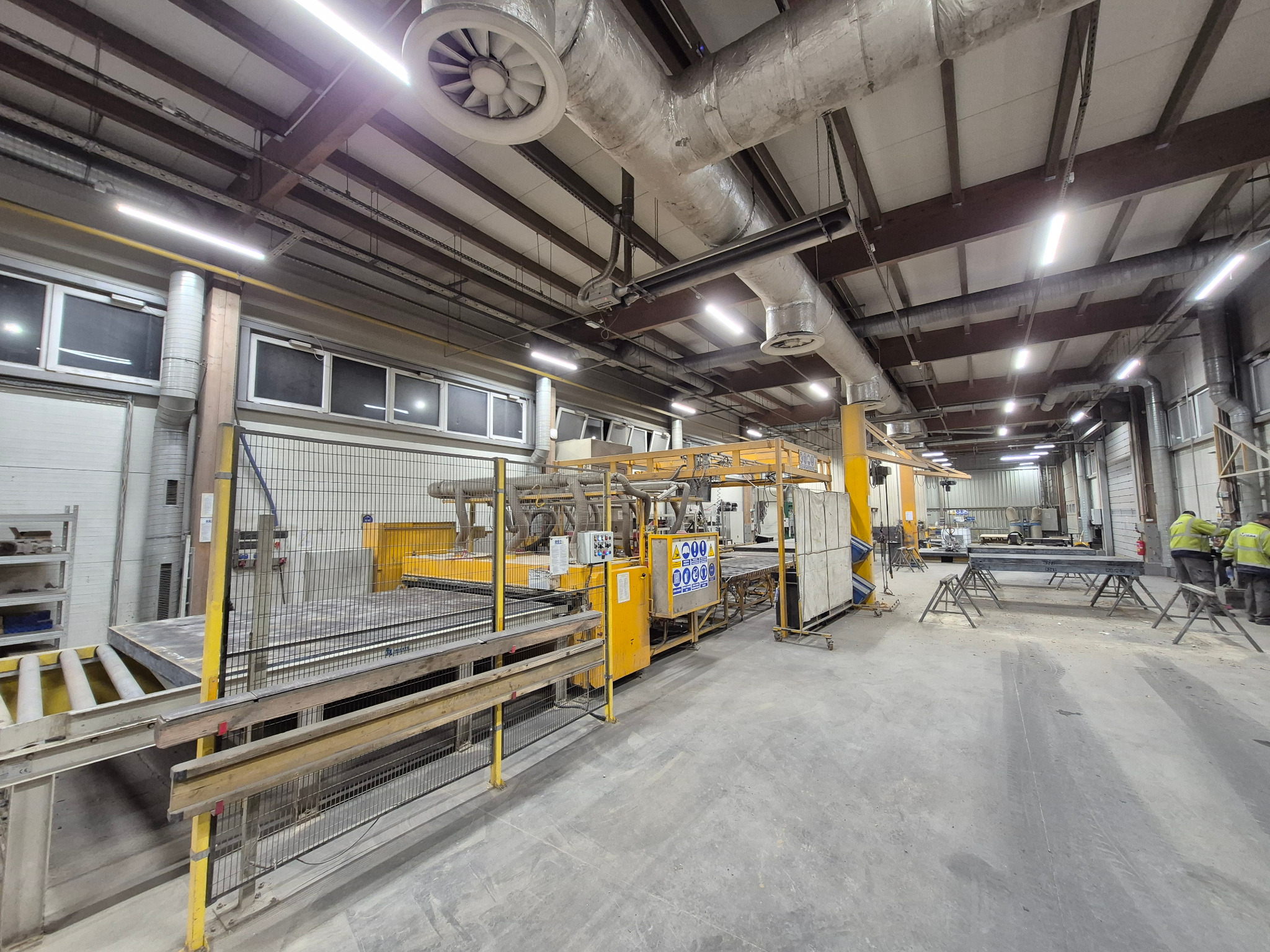 Wnętrze hali produkcyjnej z systemem oświetlenia jarzeniowego zamontowanym pod sufitem, widoczne instalacje wentylacyjne i maszyny produkcyjne.