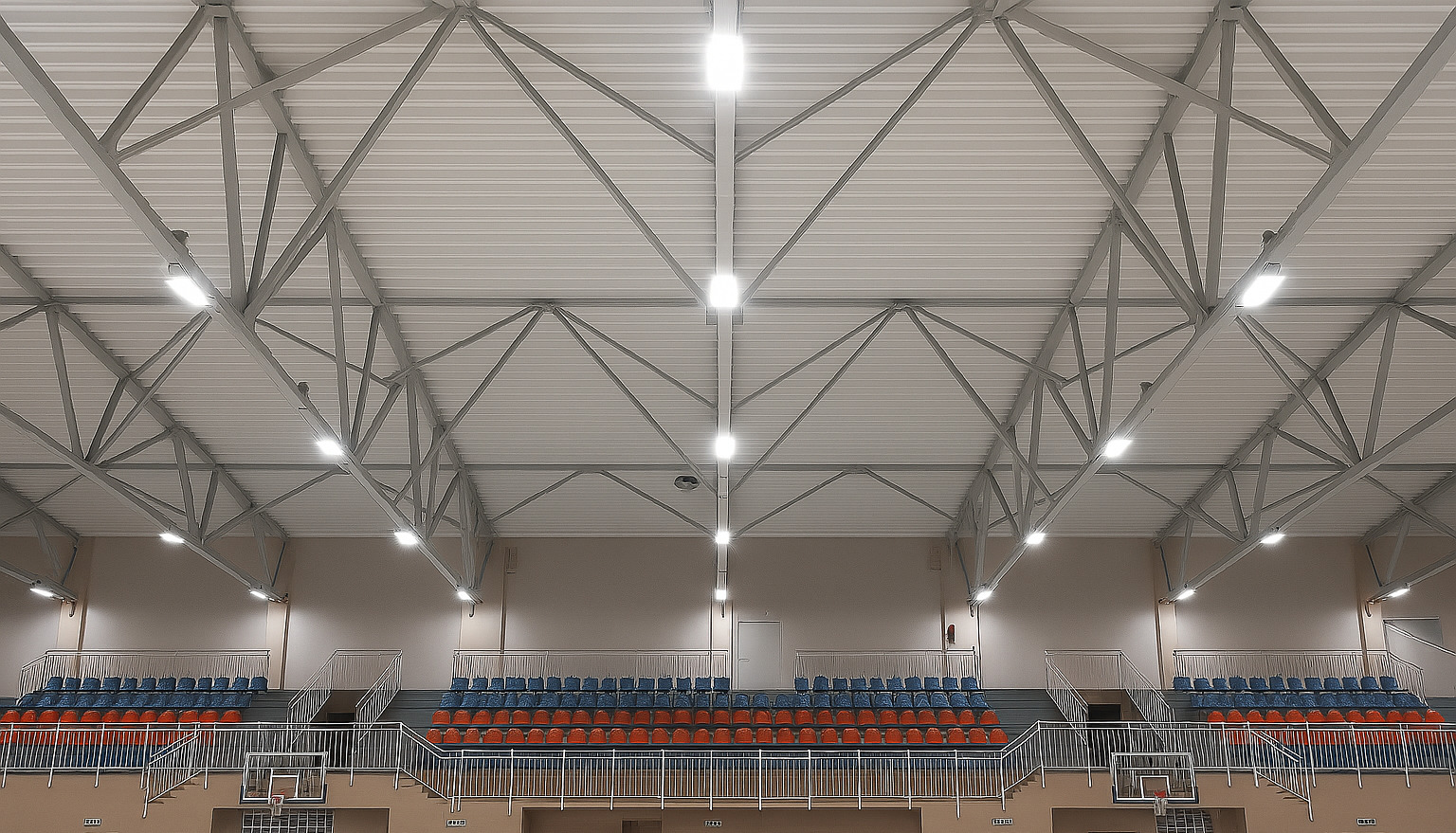 Hala sportowa z widoczną konstrukcją dachu i rzędami krzeseł. Zamontowane, równomiernie rozmieszczone lampy sufitowe oświetlają przestrzeń.