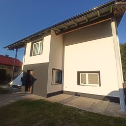 RPGO SPÓŁKA Z OGRANICZONĄ ODPOWIEDZIALNOŚCIĄ - Dwukondygnacyjny dom z elewacją w odcieniach bieli i szarości, z widocznymi oknami z żaluzjami, na tle błękitnego nieba i zieleni. Ujęcie z dołu.