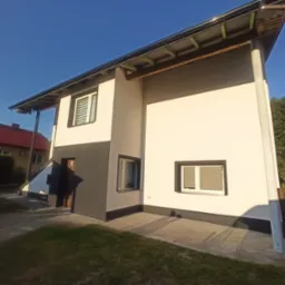 Dwukondygnacyjny dom z elewacją w odcieniach bieli i szarości, z widocznymi oknami z żaluzjami, na tle błękitnego nieba i zieleni. Ujęcie z dołu.