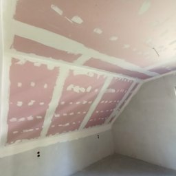RPGO SPÓŁKA Z OGRANICZONĄ ODPOWIEDZIALNOŚCIĄ - Poddasze z zamontowaną płytą karton-gips, widoczne łączenia płyt przygotowane do szpachlowania. Ściany i podłoga surowe, w odcieniach szarości.
