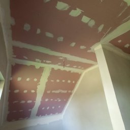 RPGO SPÓŁKA Z OGRANICZONĄ ODPOWIEDZIALNOŚCIĄ - Skośny sufit z płyt kartonowo-gipsowych w trakcie szpachlowania, widoczne łączenia i miejsca po wkrętach. Ściany w jasnym odcieniu, sufit w kolorze różowym.
