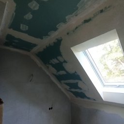 RPGO SPÓŁKA Z OGRANICZONĄ ODPOWIEDZIALNOŚCIĄ - Poddasze z oknem dachowym w trakcie prac wykończeniowych: szpachlowanie łączeń płyt gipsowo-kartonowych na suficie i ścianach. Widoczne niedoskonałości i surowy stan.