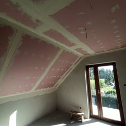 RPGO SPÓŁKA Z OGRANICZONĄ ODPOWIEDZIALNOŚCIĄ - Poddasze ze skośnym sufitem, płyty karton-gips na konstrukcji, widoczne łączenia płyt, przygotowane do szpachlowania. Okno balkonowe wpuszcza jasne światło.