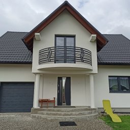 RPGO SPÓŁKA Z OGRANICZONĄ ODPOWIEDZIALNOŚCIĄ - Nowoczesny dom jednorodzinny z balkonem i grafitową stolarką okienną. Biała elewacja, ciemna dachówka, minimalistyczne detale. Widok z przodu, pochmurne niebo.