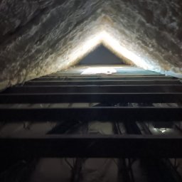 RPGO SPÓŁKA Z OGRANICZONĄ ODPOWIEDZIALNOŚCIĄ - Poddasze z widoczną izolacją pianką PUR, ujęcie od dołu. Widoczne drewniane belki konstrukcyjne i szczeliny między nimi. Izolacja w kolorze jasnobeżowym.