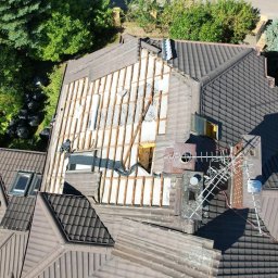 RPGO SPÓŁKA Z OGRANICZONĄ ODPOWIEDZIALNOŚCIĄ - Demontaż starego pokrycia dachowego z widoczną konstrukcją i ociepleniem. Prace dekarskie w toku, widok z drona na brązowy dach i czarne worki na odpady.