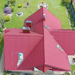 RPGO SPÓŁKA Z OGRANICZONĄ ODPOWIEDZIALNOŚCIĄ - Czerwony dach domu z blachodachówki, widok z góry na tle zielonego ogrodu. Widoczne kominy i okno dachowe. Ujęcie z drona.