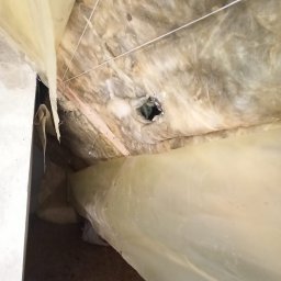 RPGO SPÓŁKA Z OGRANICZONĄ ODPOWIEDZIALNOŚCIĄ - Ocieplenie poddasza wełną mineralną, zabezpieczone folią paroizolacyjną. Widoczny otwór w izolacji i drewniana konstrukcja dachu. Ujęcie z dołu, w trakcie prac.