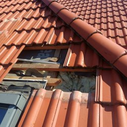 RPGO SPÓŁKA Z OGRANICZONĄ ODPOWIEDZIALNOŚCIĄ - Czerwona dachówka z widocznym oknem dachowym i izolacją z wełny mineralnej. Fragment dachu z uszkodzeniami i elementami drewnianymi. Prace dekarskie w toku.