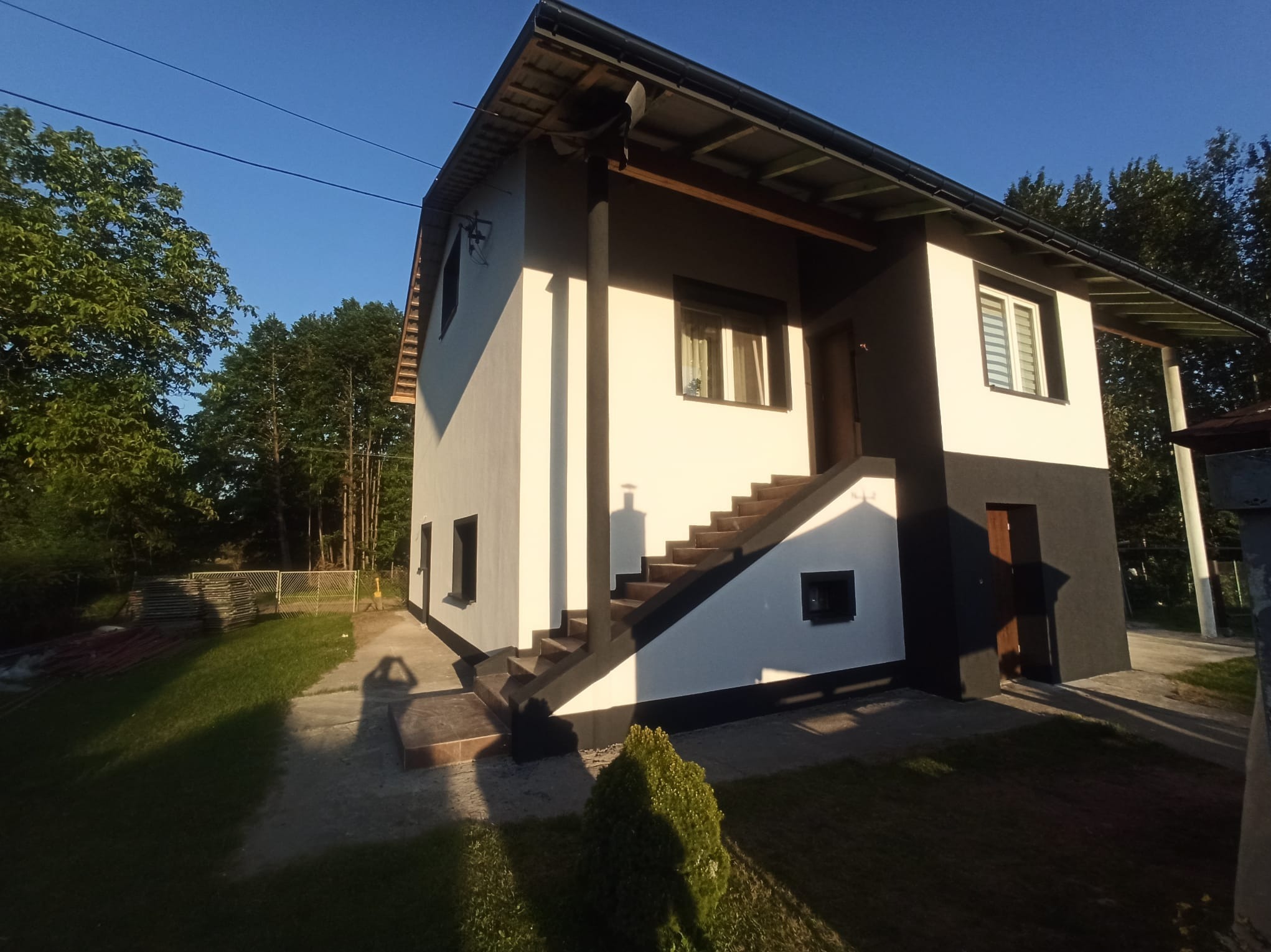 Dwukondygnacyjny dom z elewacją w bieli i antracycie, schody zewnętrzne prowadzące do drzwi, zieleń w otoczeniu, cień osoby fotografującej.