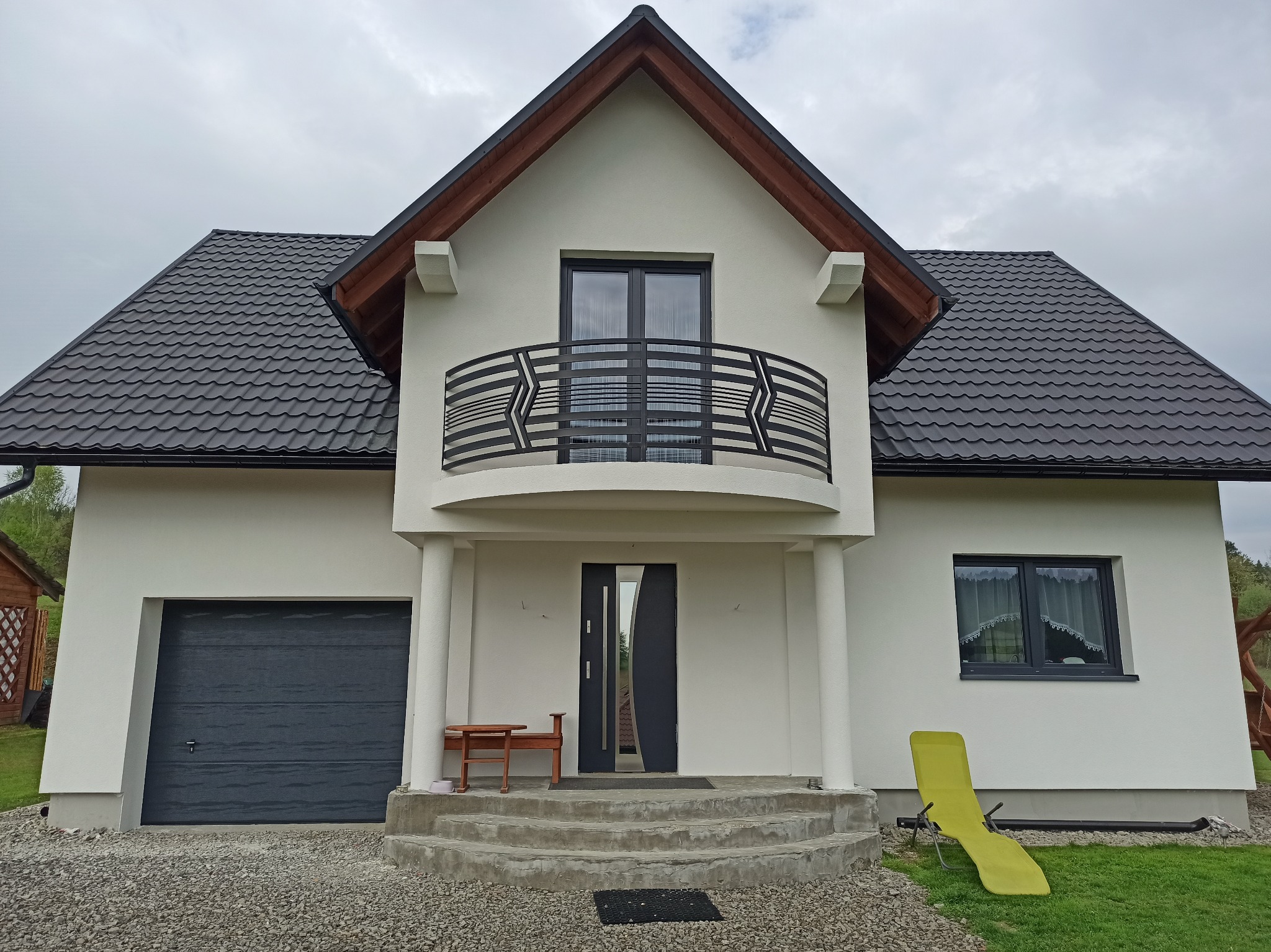 Nowoczesny dom jednorodzinny z balkonem i grafitową stolarką okienną. Biała elewacja, ciemna dachówka, minimalistyczne detale. Widok z przodu, pochmurne niebo.