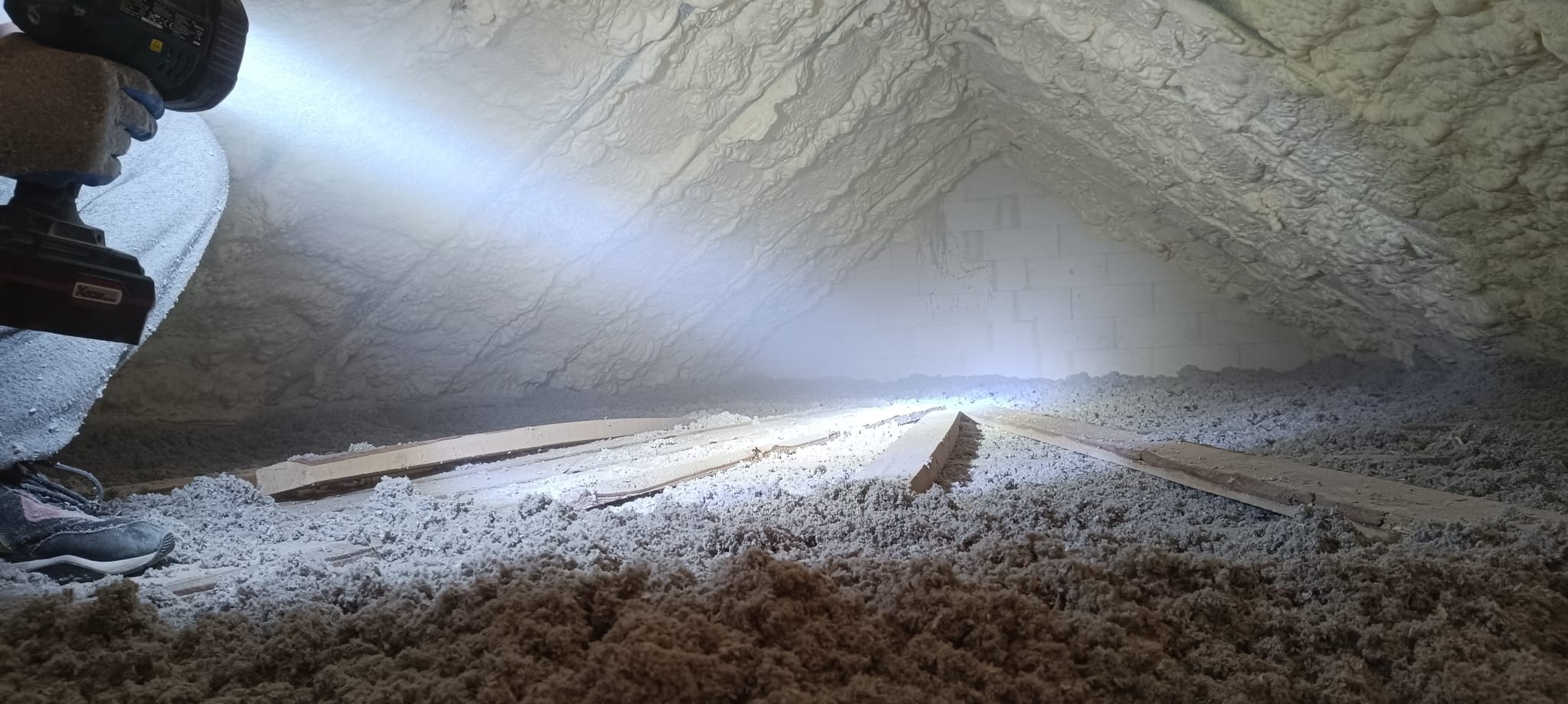 Poddasze po ociepleniu pianką PUR w Rzeszowie. Widoczna warstwa izolacji na skośnych ścianach i podłodze, oświetlona lampą. Fragment buta i narzędzia w kadrze.